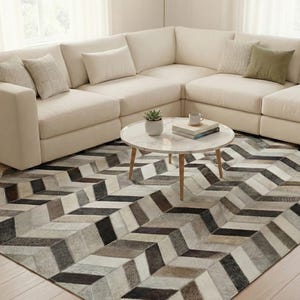 Op de afbeelding: Een groot rechthoekig vloerkleed met een herhalend visgraatpatroon in tinten zwart, grijs, wit en beige. Het kleed ligt op een lichte houten vloer voor een grote crèmekleurige hoekbank met verschillende sierkussens.