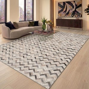 Könnte beinhalten: Ein rechteckiger Teppich mit einem geometrischen Fischgrätenmuster aus einzelnen Fellstücken in verschiedenen Nuancen von Creme, Beige, Grau und Schwarz, der eine strukturierte, mehrfarbige Oberfläche auf dem hellen Holzboden bildet.