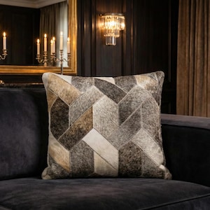 May include: Square cushion with a geometric pattern made from patches of brown, grey, and cream-coloured animal hide. The cushion sits on a dark grey sofa, offering a contrast in textures and colours.