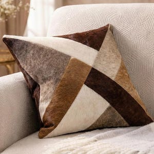 May include: A decorative square pillow with a patchwork design. The pillow features geometric shapes in shades of brown, beige, and white. The pillow is made of a soft, textured material and is placed on a neutral-colored sofa.