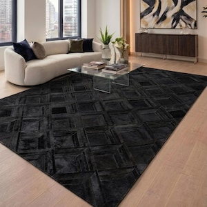 May include: A large, rectangular black rug with a geometric cube pattern dominates the foreground. The rug is placed in a modern living room with a white sofa, glass coffee table, and a dark wood cabinet. The room has large windows.
