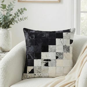 Puede incluir: Cojín decorativo cuadrado con un diseño de patchwork en piel de vaca negra, blanca y gris. El cojín está colocado sobre un sillón blanco con una manta color crema. Un jarrón con vegetación está en el fondo.