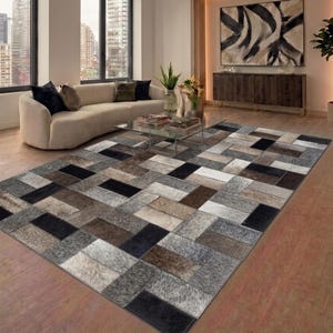 Puede incluir: Una alfombra rectangular con un diseño de patchwork. La alfombra presenta parches rectangulares en tonos de negro, gris, marrón y crema. La alfombra está colocada sobre un suelo de madera en un salón.