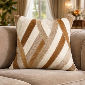 May include: Square cushion with a geometric pattern. The cushion has a patchwork design with strips of brown, tan, and cream-coloured leather or hide. It is placed on a beige sofa, suggesting a home decor theme.