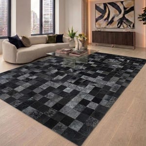 Op de afbeelding: Een groot, rechthoekig vloerkleed met een patchwork-ontwerp in tinten zwart, grijs en houtskool. Het kleed bestaat uit vierkanten met verschillende texturen en tinten, waardoor een geometrisch patroon ontstaat. Het ligt op een lichtgekleurde houten vloer.