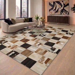 May include: A large, rectangular rug with a patchwork design. The rug features squares of various shades of brown, beige, and black, creating a geometric pattern. The rug is placed on a light-coloured wooden floor.