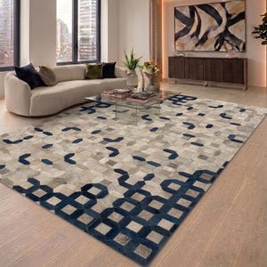 Op de afbeelding: Een groot vloerkleed met een geometrisch patroon in grijstinten en marineblauw. Het kleed is gemaakt van een patchwork van vormen, wat een modern design creëert. Het ligt op een lichtgekleurde houten vloer.