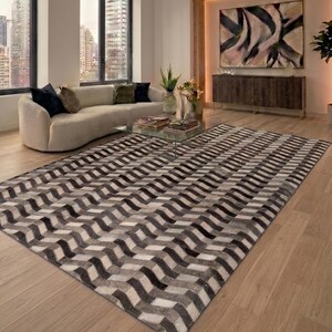 Op de afbeelding: Een moderne woonkamer met een groot vloerkleed met een geometrisch zigzagpatroon in grijstinten en wit. Het vloerkleed ligt op een lichte houten vloer, als aanvulling op de neutrale tinten van de meubels en de inrichting van de kamer.