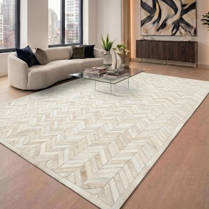 May include: A large, rectangular area rug with a herringbone pattern in shades of cream and beige. The rug is placed on a hardwood floor in a modern living room setting. The room also features a curved sofa, glass coffee table, and abstract artwork.