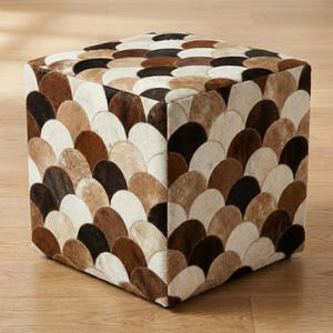 May include: A square ottoman with a cowhide patchwork design. The ottoman features a repeating pattern of fan-shaped pieces in shades of brown, beige, and black. The overall aesthetic is rustic and textured, suitable for living room decor.