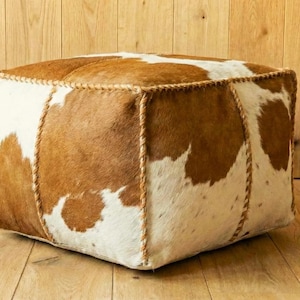 Könnte beinhalten: Quadratischer Hocker mit einem Kuhfellmuster in Braun- und Weißtönen. Die Kanten sind mit hellbraunem Faden vernäht. Dieses dekorative Möbelstück eignet sich für ein Wohn- oder Schlafzimmer. Ca. 60 cm x 60 cm.