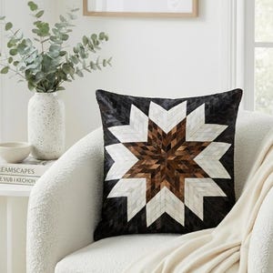 Op de afbeelding: Een vierkant decoratief kussen met een sterrenpatroon. Het kussen heeft een zwarte achtergrond met een geometrisch sterrenontwerp in wit, bruin en beige. Het ligt op een witte fauteuil, met een crèmekleurige deken.