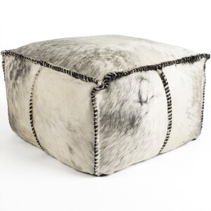 May include: Square ottoman with a cowhide exterior in shades of gray and white. The edges are accented with black stitching, creating a textured and stylish piece of furniture. The ottoman is a cube shape.
