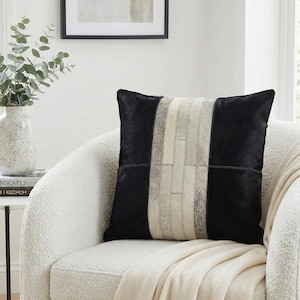 Könnte beinhalten: Ein quadratisches Zierkissen mit einem Patchwork-Design aus schwarzem und cremefarbenem Fell. Das Kissen hat zwei einfarbige schwarze Seitenteile und einen vertikalen Mittelstreifen aus kleineren, rechteckigen Stücken aus cremefarbenem und grauem Fell.