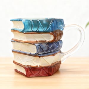 May include: A novelty glass mug designed as a stack of books. The mug has a clear handle and the books are in blue, brown, and red. The pages are white, and the mug is on a wooden surface.