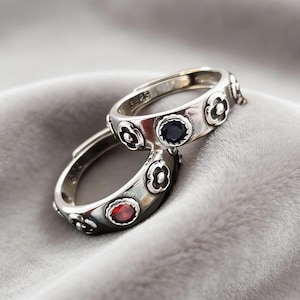 May include: Two silver rings with floral patterns and adjustable bands. One ring has a red gemstone, the other a dark blue gemstone. The rings are displayed on a soft, grey background.