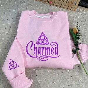 May include: A light pink crewneck sweatshirt featuring the word Charmed in purple embroidery across the chest, accompanied by a purple triquetra symbol above the text and a matching purple triquetra embroidered on the left sleeve cuff.