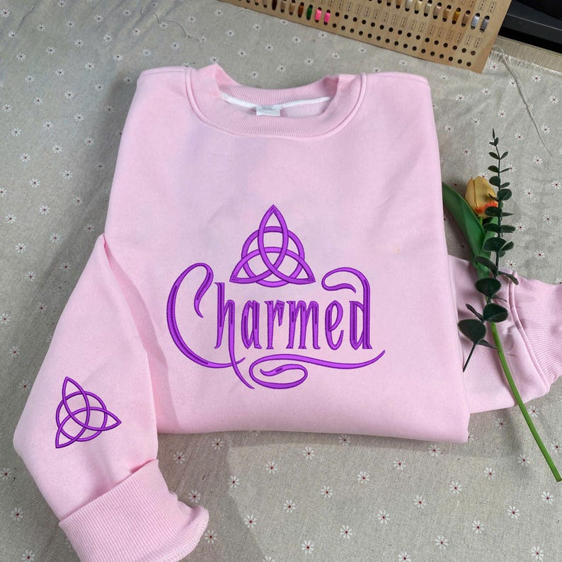 May include: A light pink crewneck sweatshirt featuring the word Charmed in purple embroidery across the chest, accompanied by a purple triquetra symbol above the text and a matching purple triquetra embroidered on the left sleeve cuff.