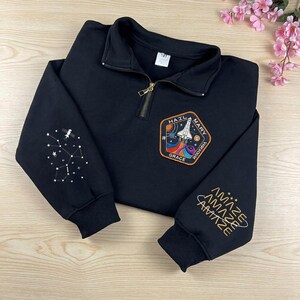 Puede incluir: Una sudadera negra con cremallera de cuarto de largo y un parche temático espacial en el pecho con un transbordador, el texto HAIL MARY GRACE y una estela colorida. Las mangas muestran constelaciones blancas bordadas y la palabra AMAZE en dorado.