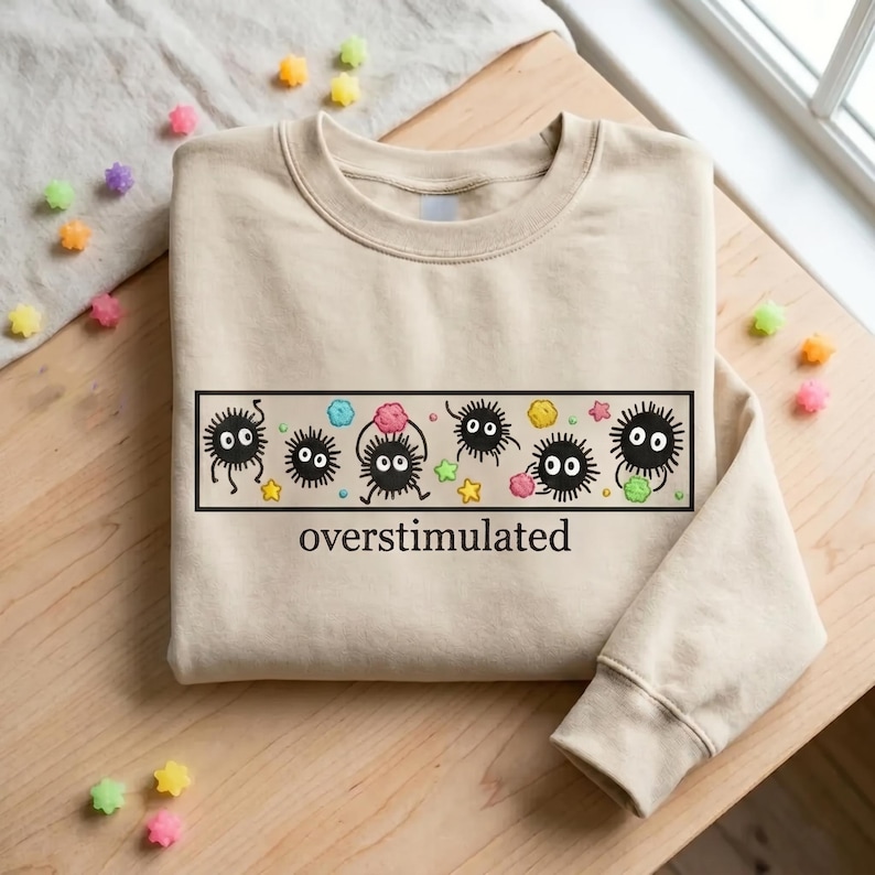 May include: Beige sweatshirt featuring a black rectangular graphic with cartoon soot sprites, colorful accents, and the word "overstimulated" in black. The design includes stars and colorful candy-like shapes.