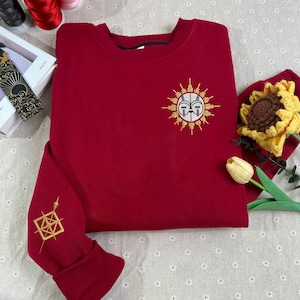Puede incluir: Una sudadera burdeos con diseños dorados bordados. La parte delantera presenta un emblema solar con una cara, y la manga izquierda tiene un patrón geométrico. La sudadera se muestra sobre una superficie con estampado floral.