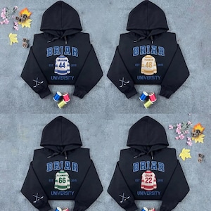 Könnte beinhalten: Vier schwarze Kapuzenpullover mit dem Wort "BRIAR" in blauen Buchstaben. Jeder Pullover hat eine andere Nummer und einen anderen Namen auf der Vorderseite sowie das Wort "UNIVERSITY". Hockeyschläger sind auf den Ärmeln.