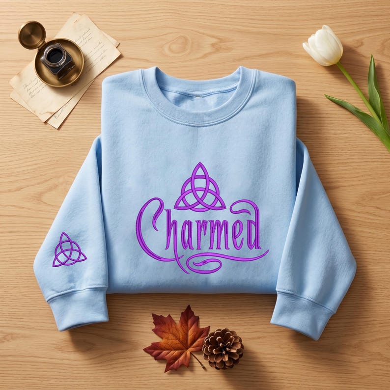 May include: A light blue crewneck sweatshirt featuring the word Charmed in purple script lettering across the chest, accompanied by a purple triquetra symbol above the text and a smaller matching triquetra symbol on the right sleeve.