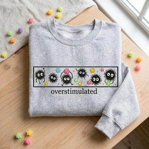 May include: A light gray sweatshirt featuring a rectangular graphic with cartoon soot sprites and the word "overstimulated" in black text. The graphic includes colorful stars and other shapes. The sweatshirt is on a wooden surface.