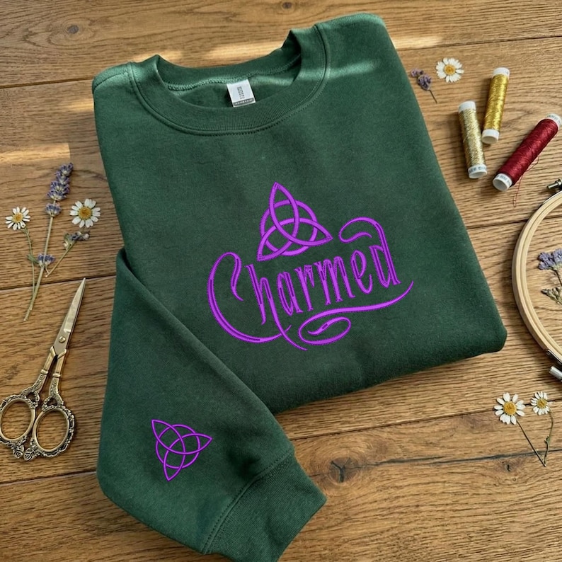 May include: A dark green crewneck sweatshirt featuring the word Charmed in purple script lettering with a purple triquetra symbol above the text and a smaller matching triquetra symbol embroidered on the sleeve cuff of the garment.