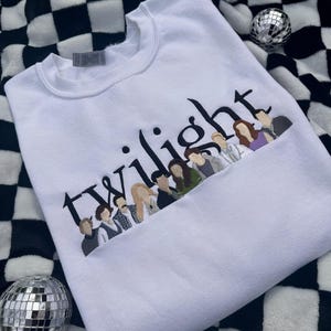 May include: White sweatshirt with the word "Twilight" embroidered in black, with a line of character illustrations below. The sweatshirt is made of a soft material. The background is a black and white checkered pattern.
