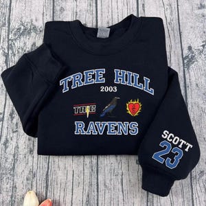 May include: Black sweatshirt with the text "TREE HILL 2003 RAVENS" in white and blue. Features a raven graphic and the name "SCOTT 23" on the sleeve. The sweatshirt is folded on a wooden surface.