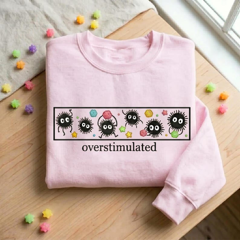 May include: A light pink sweatshirt with a black rectangular graphic featuring cartoon soot sprites and the word "overstimulated". The graphic includes colorful stars and gem-like shapes. The sweatshirt is on a wooden surface.