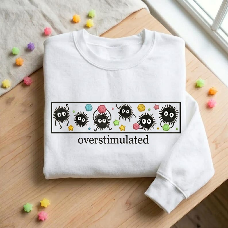 May include: White sweatshirt featuring a rectangular graphic with cartoonish black creatures and the word "overstimulated". The graphic includes colorful stars and shapes. The sweatshirt is on a wooden surface.