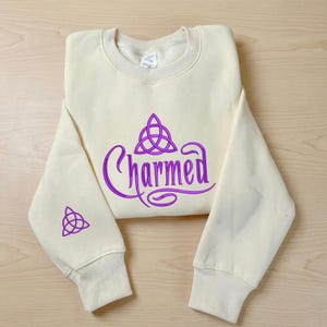 May include: A cream-colored crewneck sweatshirt featuring the word Charmed in purple script lettering with a purple triquetra symbol above it. A smaller purple triquetra symbol is printed on the lower portion of the left sleeve.