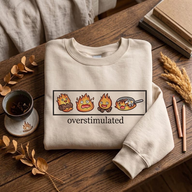 May include: Beige sweatshirt with a black rectangular graphic featuring four cartoon flames, each with a different expression. The word "overstimulated" is printed in black below the graphic. The sweatshirt is on a wooden surface.