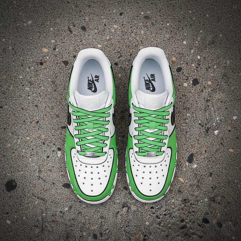 Puede incluir: Par de zapatillas Nike Air Force 1 blancas y verdes. Las zapatillas tienen cordones verdes, un contorno verde y una base blanca con peque&ntilde;as perforaciones. El logotipo de Nike Air es visible en la leng&uuml;eta y la plantilla.
