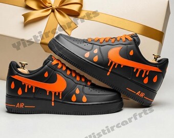 May include: Pair of black sneakers with orange accents, including the Nike swoosh and dripping paint design. The shoes have orange laces and the word "AIR" printed on the side. A gift box with a gold bow is in the background.