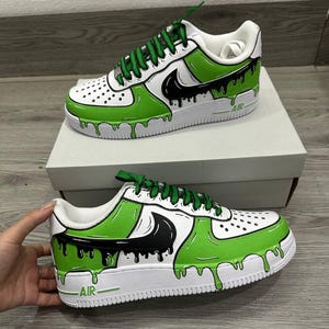 Puede incluir: Zapatillas personalizadas blancas y verdes con detalles en negro. Los zapatos presentan un dise&ntilde;o &uacute;nico con un efecto de pintura verde goteando, un logotipo negro y cordones verdes. La palabra "AIR" est&aacute; impresa en el lateral de la suela blanca.