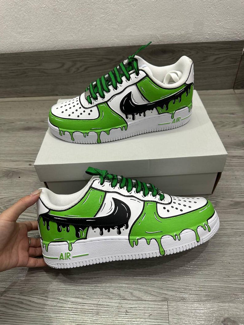 Puede incluir: Zapatillas personalizadas blancas y verdes con detalles en negro. Los zapatos presentan un dise&ntilde;o &uacute;nico con un efecto de pintura verde goteando, un logotipo negro y cordones verdes. La palabra "AIR" est&aacute; impresa en el lateral de la suela blanca.