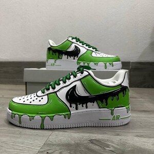 Puede incluir: Zapatillas blancas y verdes con detalles en negro. Los zapatos presentan un dise&ntilde;o &uacute;nico con pintura verde goteando por los lados y un logotipo de swoosh negro. Los cordones son verdes y la palabra "AIR" es visible en la suela.