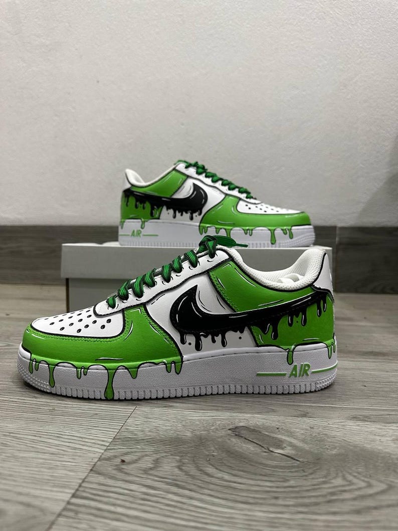 Puede incluir: Zapatillas blancas y verdes con detalles en negro. Los zapatos presentan un dise&ntilde;o &uacute;nico con pintura verde goteando por los lados y un logotipo de swoosh negro. Los cordones son verdes y la palabra "AIR" es visible en la suela.