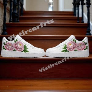 May include: White sneakers with a floral design. The shoes feature pink peonies with green leaves painted on the sides. The sneakers have a white sole and laces.