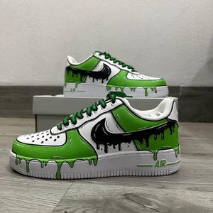 Puede incluir: Zapatillas blancas y verdes con detalles negros y un dise&ntilde;o de pintura goteando. Las zapatillas presentan un logotipo de Nike negro y cordones verdes. La palabra "AIR" est&aacute; impresa en el lateral de la suela blanca.