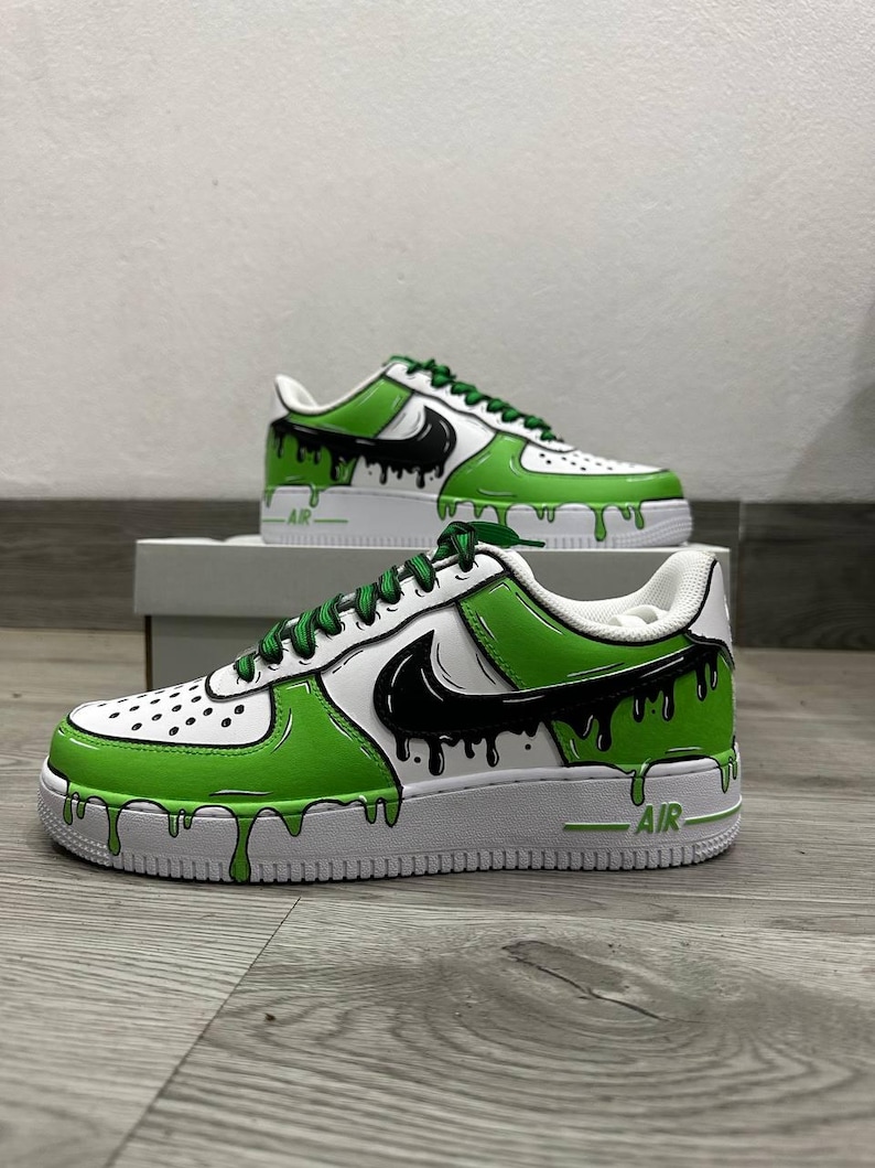 Puede incluir: Zapatillas blancas y verdes con detalles negros y un dise&ntilde;o de pintura goteando. Las zapatillas presentan un logotipo de Nike negro y cordones verdes. La palabra "AIR" est&aacute; impresa en el lateral de la suela blanca.