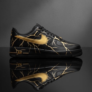 May include: Black and gold athletic shoe with a lightning bolt design. The shoe features a black base with gold accents, including the Nike swoosh and lightning bolt patterns. The word "AIR" is printed on the side.