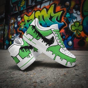 Puede incluir: Zapatillas blancas y verdes con un dise&ntilde;o de goteo &uacute;nico. Las zapatillas presentan un logotipo negro y cordones verdes. La palabra "AIR" es visible en el lateral. Las zapatillas est&aacute;n sobre un fondo de graffiti colorido.