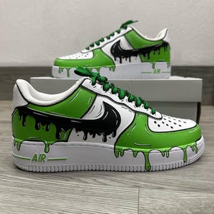 Puede incluir: Zapatillas blancas personalizadas con detalles en verde y negro. Los zapatos presentan un dise&ntilde;o de goteo verde y negro, un logotipo de Nike negro y cordones verdes. La palabra "AIR" es visible en la suela blanca.