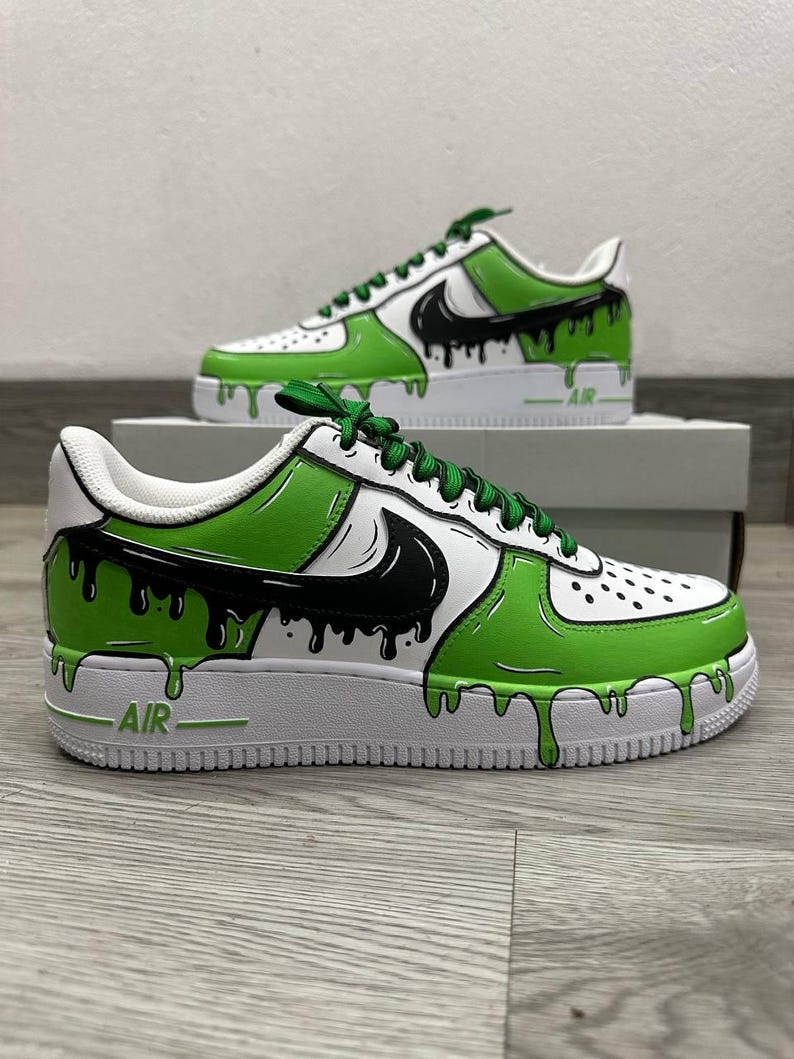 Puede incluir: Zapatillas blancas personalizadas con detalles en verde y negro. Los zapatos presentan un dise&ntilde;o de goteo verde y negro, un logotipo de Nike negro y cordones verdes. La palabra "AIR" es visible en la suela blanca.