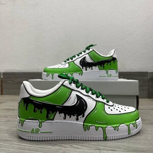 Puede incluir: Zapatillas blancas personalizadas con un dise&ntilde;o de goteo de pintura verde y negra. Las zapatillas presentan un logotipo negro, cordones verdes y la palabra "AIR" en el lateral. El dise&ntilde;o est&aacute; sobre una suela blanca.