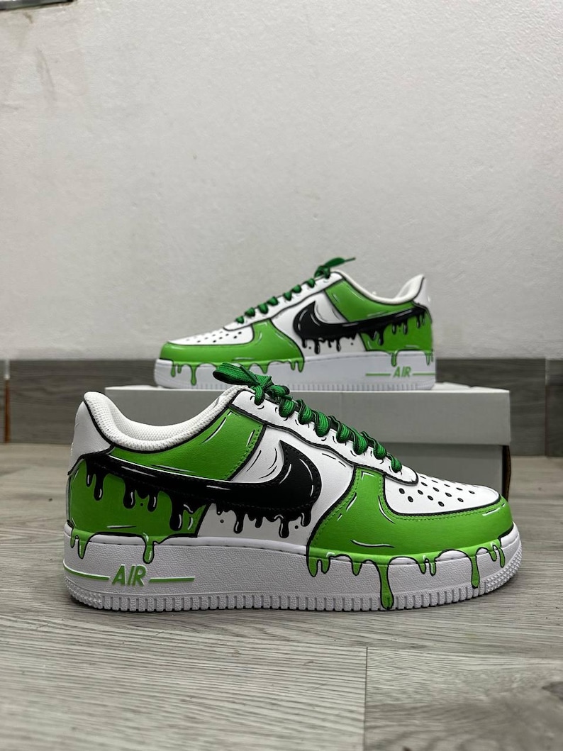 Puede incluir: Zapatillas blancas personalizadas con un dise&ntilde;o de goteo de pintura verde y negra. Las zapatillas presentan un logotipo negro, cordones verdes y la palabra "AIR" en el lateral. El dise&ntilde;o est&aacute; sobre una suela blanca.